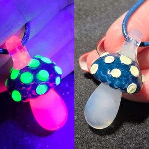 Split image of the same handmade borosilicate glass mushroom pendant shown in regular light and under UV reactive black light. In normal light, the mushroom has a blue frit cap with pale yellow raised dots and a soft milky white stem. Under black light, the pendant dramatically color shifts with neon green glowing dots, a bright pink glowing stem, and a pink-purple cap, highlighting the UV reactive glass details.