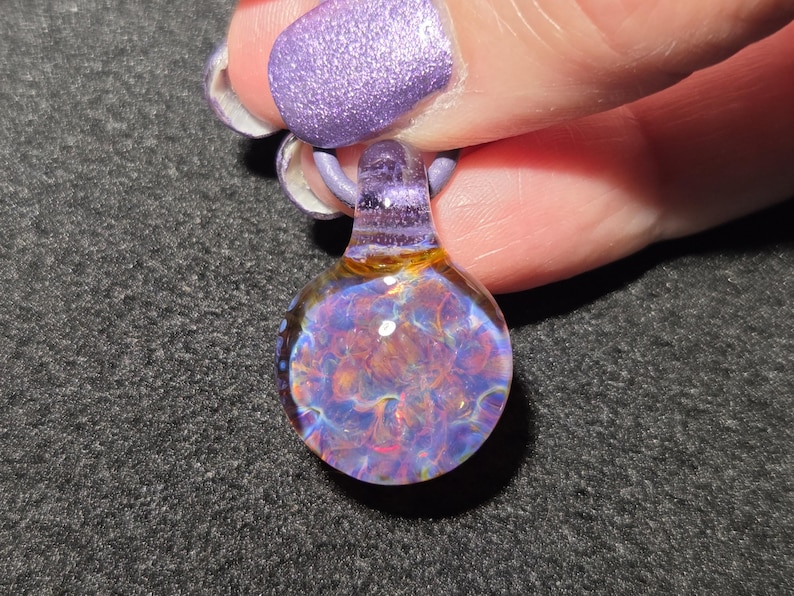 Small hand blown borosilicate implosion pendant with layered purple, blue, pink and golden frit, photographed against a dark background.