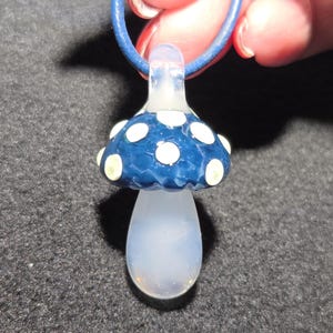 Handheld borosilicate glass magic mushroom pendant with a deep teal blue cap, pale yellow dot details, and a translucent clear stem, shown on a black leather cord in natural light.