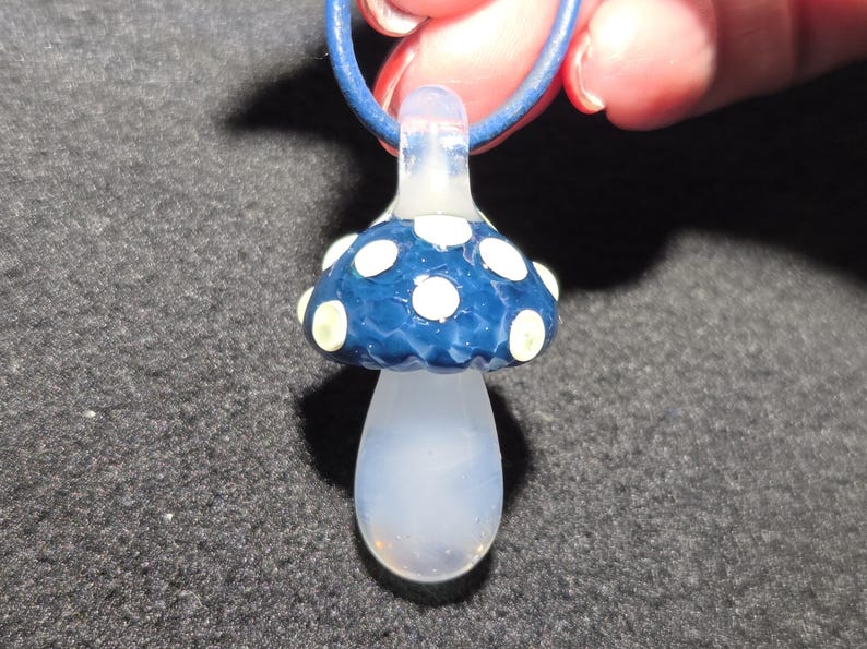 Handheld borosilicate glass magic mushroom pendant with a deep teal blue cap, pale yellow dot details, and a translucent clear stem, shown on a black leather cord in natural light.