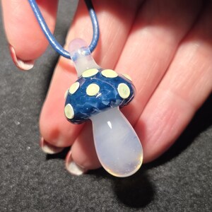 Borosilicate glass magic mushroom necklace with a glossy blue cap, creamy yellow dots, and a clear stem, displayed against a dark background without UV lighting.