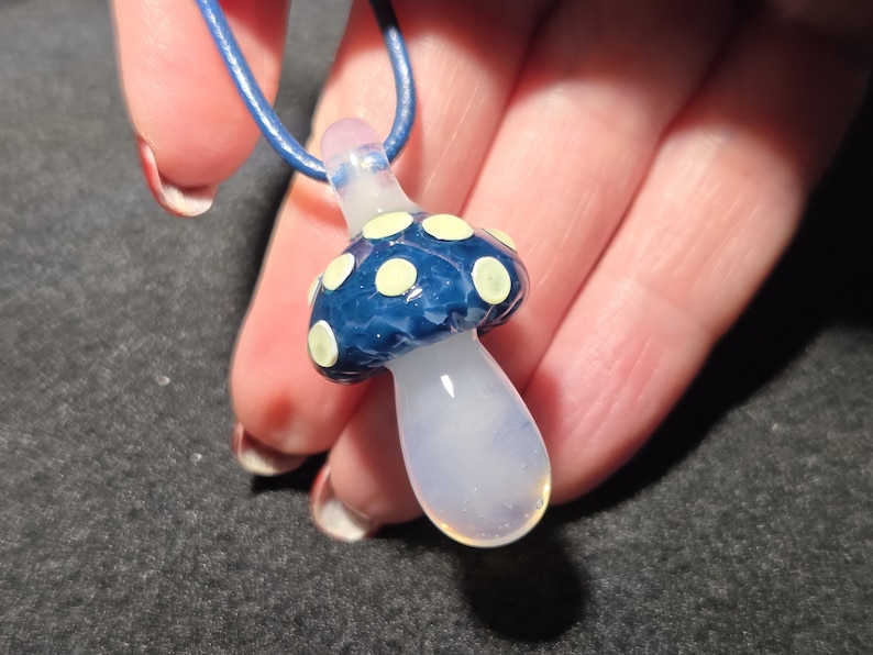 Borosilicate glass magic mushroom necklace with a glossy blue cap, creamy yellow dots, and a clear stem, displayed against a dark background without UV lighting.