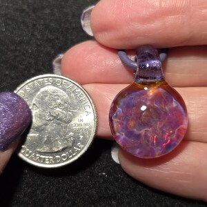 Sparkly borosilicate glass pendant featuring a deep floral implosion pattern with glowing pink, purple and blue tones and a purple glass bail.   Pendant size compared to a quarter and it is larger measuring 1" long with the glass bail.  The round portion measures .75" diameter.
