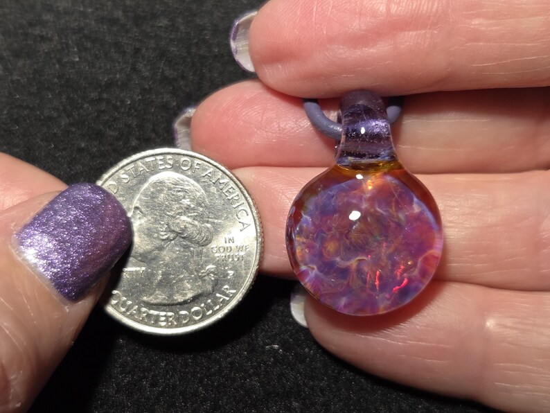 Sparkly borosilicate glass pendant featuring a deep floral implosion pattern with glowing pink, purple and blue tones and a purple glass bail.   Pendant size compared to a quarter and it is larger measuring 1" long with the glass bail.  The round portion measures .75" diameter.