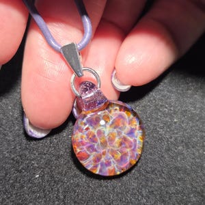 Close-up of a colorful heady glass pendant with a dainty frit implosion design in shades of violet, magenta, and amber, shown against a black background.