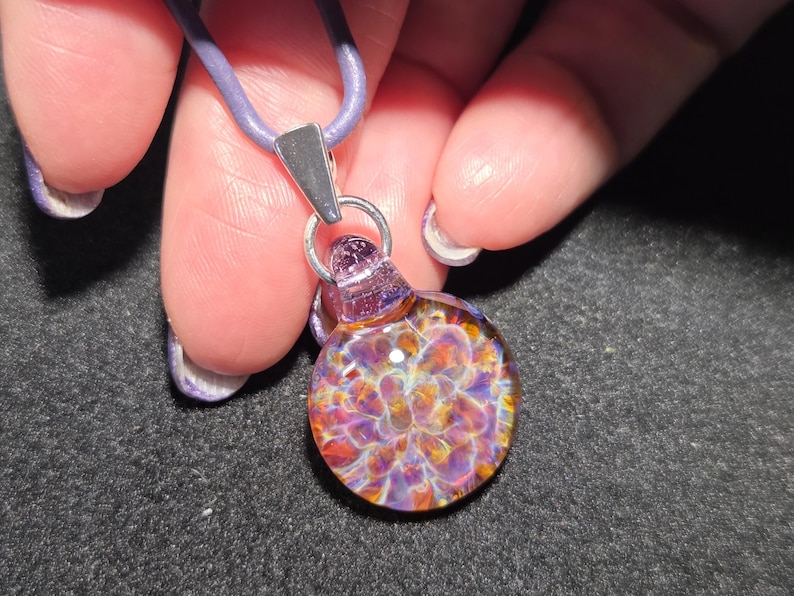 Close-up of a colorful heady glass pendant with a dainty frit implosion design in shades of violet, magenta, and amber, shown against a black background.