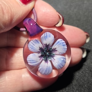 May include: A glass pendant with a floral design. The pendant features a clear, round glass base with a flower design in shades of blue, white, and purple. The pendant is held by a purple cord.
