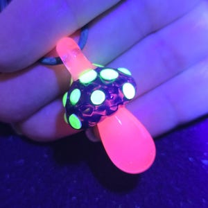 Same glass magic mushroom pendant under UV light, glowing neon green dots with a bright hot pink stem, demonstrating the color-changing reactive glass effect.