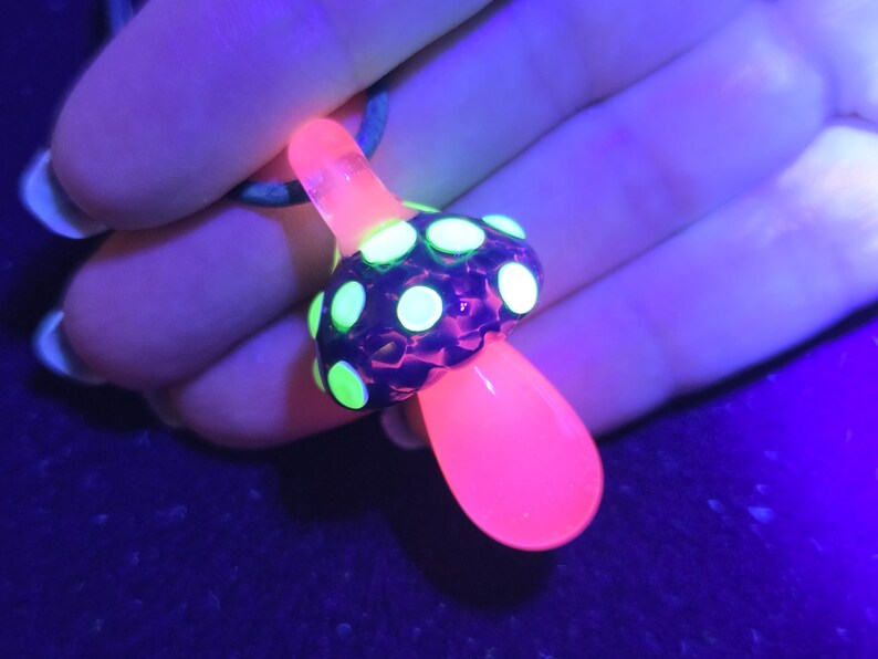 Same glass magic mushroom pendant under UV light, glowing neon green dots with a bright hot pink stem, demonstrating the color-changing reactive glass effect.