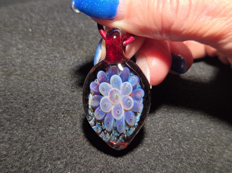 Close-up of a handcrafted glass implosion pendant with soft lavender petals layered over a rich red and blue-sparkle background, highlighting the dimensional depth and polished glass surface.