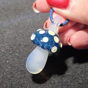 Close-up of a handmade glass mushroom pendant featuring a dark blue cap with soft yellow spots and a clear glass stem, photographed in a hand to show scale.