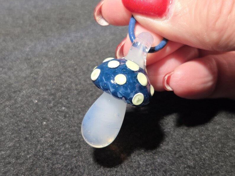 Close-up of a handmade glass mushroom pendant featuring a dark blue cap with soft yellow spots and a clear glass stem, photographed in a hand to show scale.