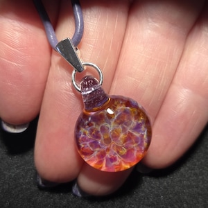 A small round blown glass implosion pendant with swirling purple, pink, and golden tones, hanging from a sterling silver bail on a leather cord, displayed in a hand.