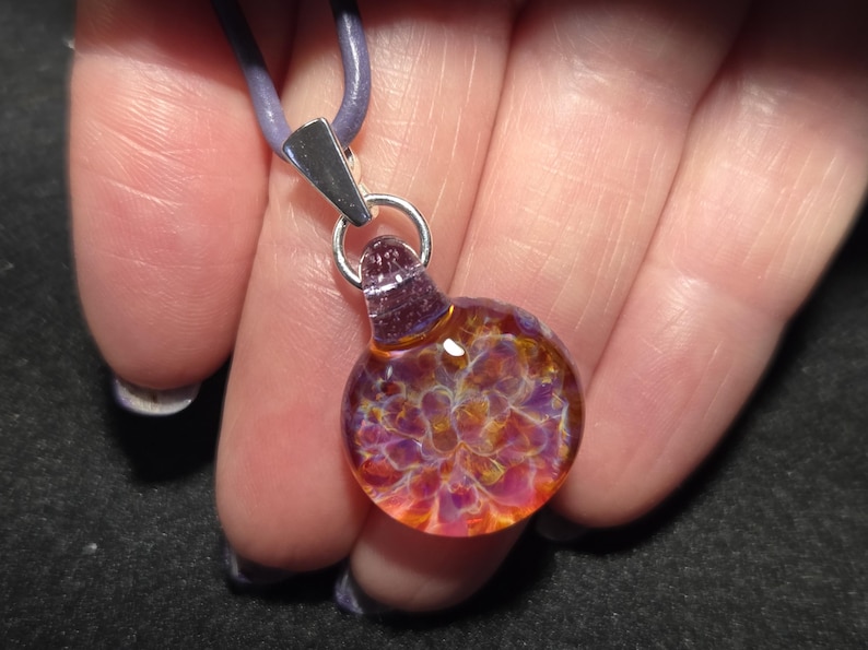 A small round blown glass implosion pendant with swirling purple, pink, and golden tones, hanging from a sterling silver bail on a leather cord, displayed in a hand.
