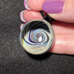 May include: A clear glass pendant with a swirling galaxy design. The pendant features a spiral of blues, purples, and whites, with a small white dot at the center. The glass is held by a black cord.