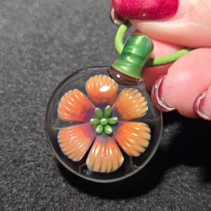 May include: A clear glass pendant with an orange and purple flower design. The flower has detailed petals and a green center. The pendant is attached to a green cord and has a green and brown glass top.
