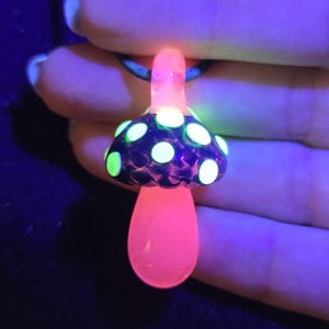 Handmade glass mushroom necklace illuminated by UV light, revealing vivid neon green spots and a bright pink stem for a psychedelic glow effect.