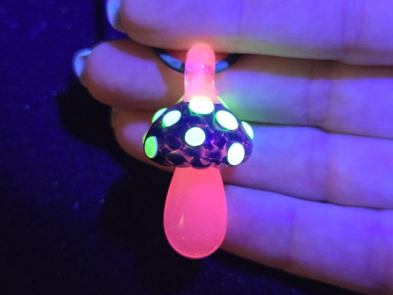 Handmade glass mushroom necklace illuminated by UV light, revealing vivid neon green spots and a bright pink stem for a psychedelic glow effect.
