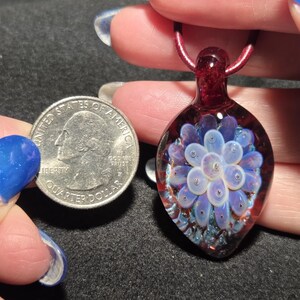 Pendant size compared to a quarter and it is larger measuring 1.75 inches long with the glass bail.  The round portion measures 1 inches diameter.  Art glass implosion pendant displaying a pale lavender flower suspended in clear borosilicate glass, surrounded by a crimson red field with shimmering blue sparkles, photographed in natural light.