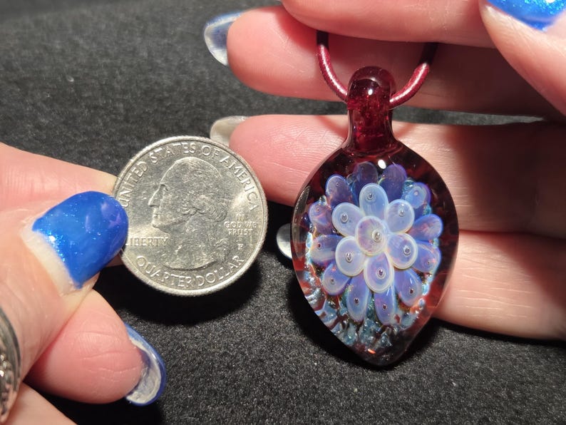 Pendant size compared to a quarter and it is larger measuring 1.75 inches long with the glass bail.  The round portion measures 1 inches diameter.  Art glass implosion pendant displaying a pale lavender flower suspended in clear borosilicate glass, surrounded by a crimson red field with shimmering blue sparkles, photographed in natural light.