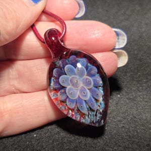 Handmade borosilicate glass implosion pendant resting on an open palm, featuring a pale lavender floral center over a deep crimson red background with blue sparkle details and a glossy glass bail.