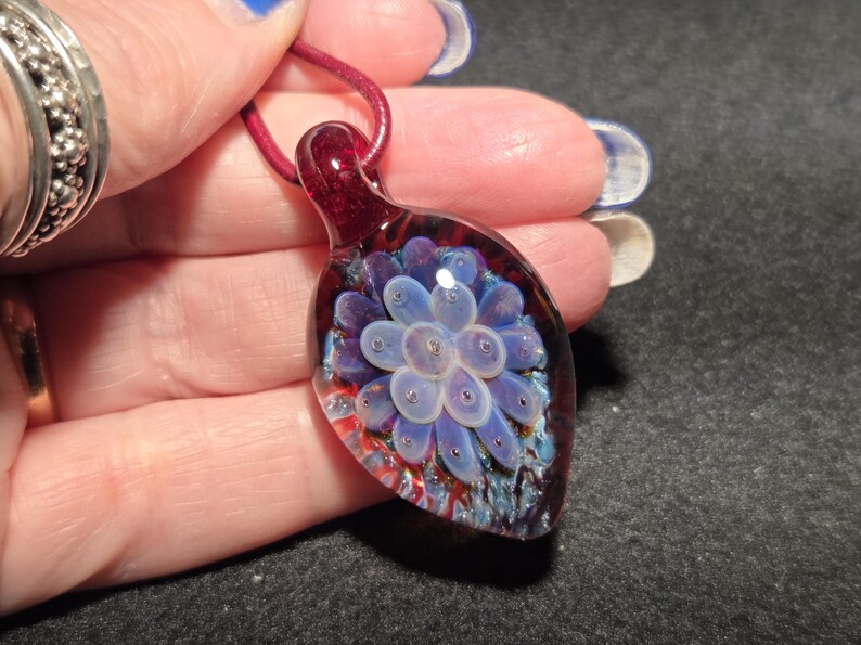 Handmade borosilicate glass implosion pendant resting on an open palm, featuring a pale lavender floral center over a deep crimson red background with blue sparkle details and a glossy glass bail.