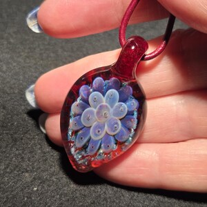 Glass implosion pendant shown on a hand for scale, with a soft lavender flower design floating within clear glass, framed by a red crimson and blue-sparkle background.