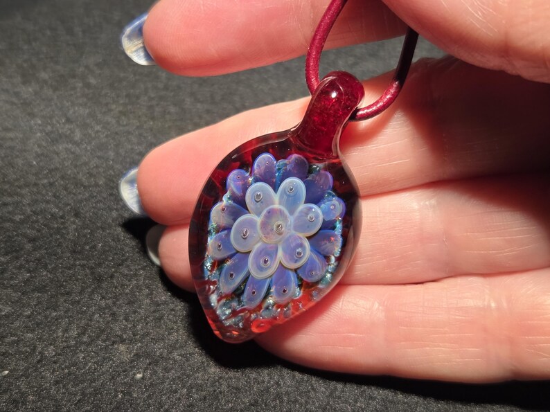 Glass implosion pendant shown on a hand for scale, with a soft lavender flower design floating within clear glass, framed by a red crimson and blue-sparkle background.