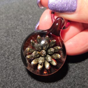Round glass pendant featuring a dimensional metallic floral design with raised golden petals and a rich red backing, shown against a dark background for contrast.