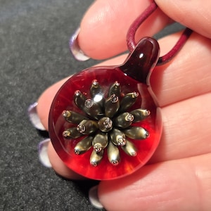 A handmade borosilicate glass pendant featuring a metallic gold and bronze implosion flower design suspended inside clear glass with a deep red background, shown resting on a hand.