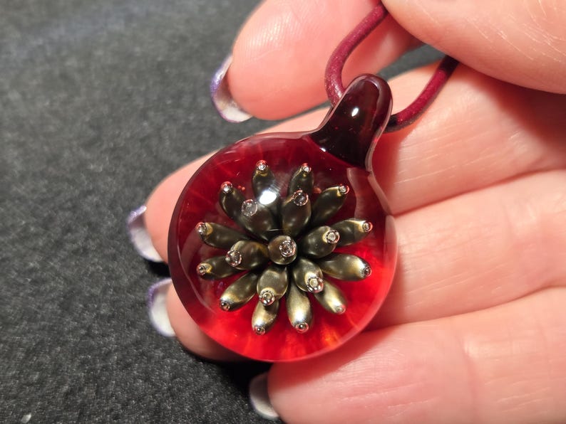 A handmade borosilicate glass pendant featuring a metallic gold and bronze implosion flower design suspended inside clear glass with a deep red background, shown resting on a hand.