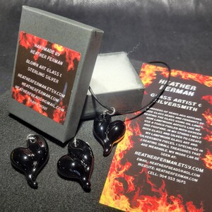 May include: A black glass heart pendant necklace with a black cord. The pendant is handmade by Heather Ferman, a glass artist and silversmith. The necklace comes in a black box with a red and black label that says "Handmade by Heather Ferman, Blown Art Glass & Sterling Silver".