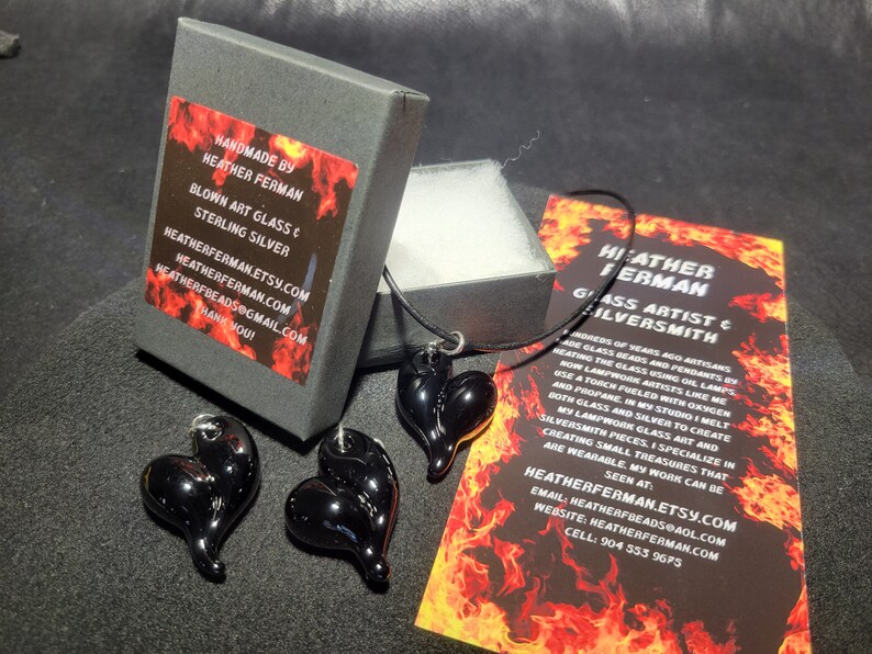 May include: A black glass heart pendant necklace with a black cord. The pendant is handmade by Heather Ferman, a glass artist and silversmith. The necklace comes in a black box with a red and black label that says "Handmade by Heather Ferman, Blown Art Glass & Sterling Silver".