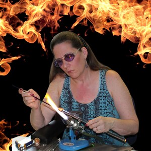 May include: A woman wearing glasses and a blue and black patterned tank top is working with a glassblowing torch. She is holding a glass rod over the flame, heating it up. The background is a fiery orange and yellow.