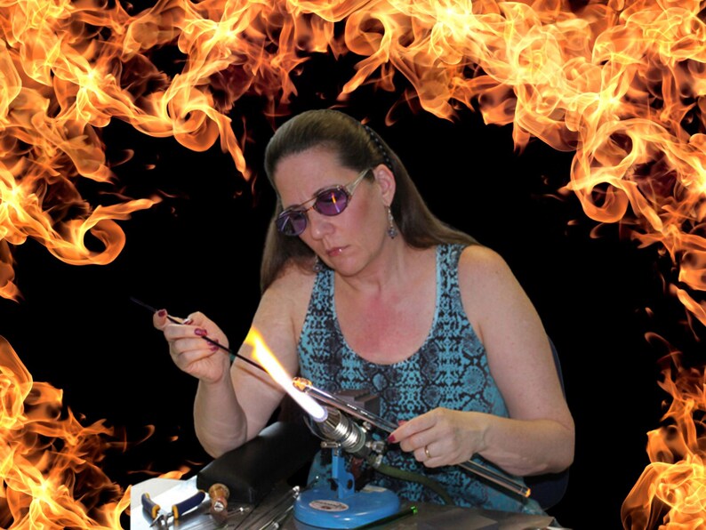 May include: A woman wearing glasses and a blue and black patterned tank top is working with a glassblowing torch. She is holding a glass rod over the flame, heating it up. The background is a fiery orange and yellow.