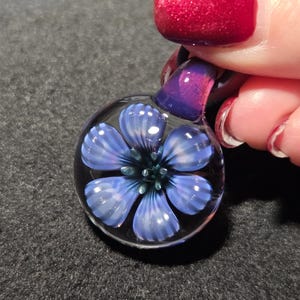 May include: A clear glass pendant with a blue flower design. The flower has five petals with white accents and a dark blue center. The pendant is held by a purple loop.