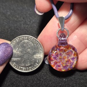 Close-up of a dainty frit pendant choker, showing intricate handcrafted glass details with a mix of fiery autumn and cool violet hues.  The pendant is pictured next to a quarter for size.