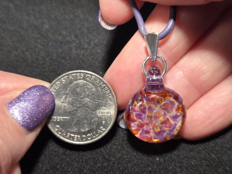 Close-up of a dainty frit pendant choker, showing intricate handcrafted glass details with a mix of fiery autumn and cool violet hues.  The pendant is pictured next to a quarter for size.