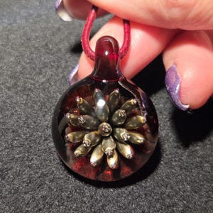 A handcrafted lampwork pendant with a 3D metallic flower implosion in gold tones, encased in clear glass with a ruby-red background, lying on a black surface to highlight its shine and depth.