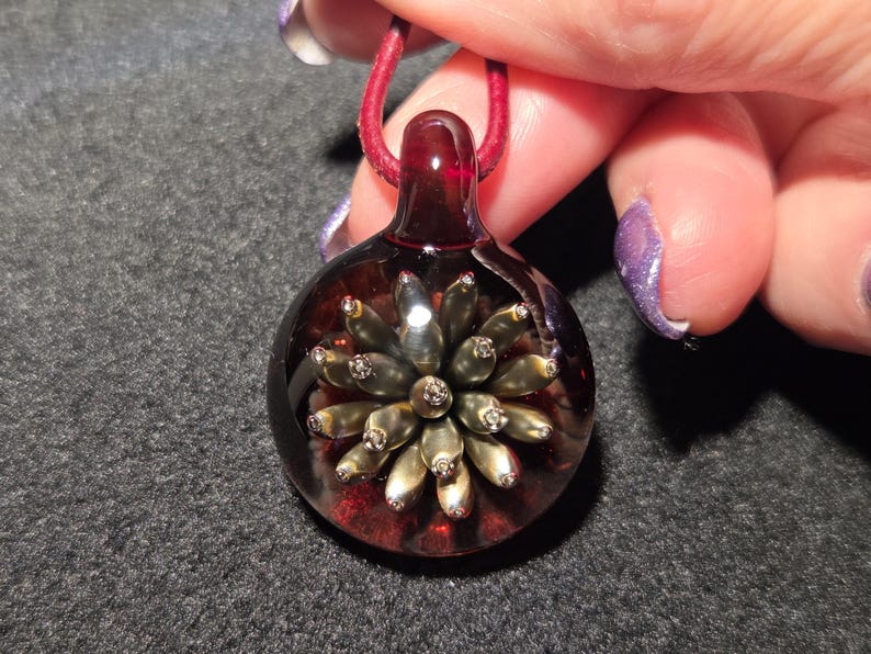 A handcrafted lampwork pendant with a 3D metallic flower implosion in gold tones, encased in clear glass with a ruby-red background, lying on a black surface to highlight its shine and depth.