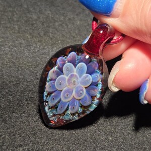 Hand-blown borosilicate glass implosion pendant featuring a pale lavender floral design encased in clear glass, set against a deep crimson red background with subtle blue sparkle accents, shown close-up against a dark surface.
