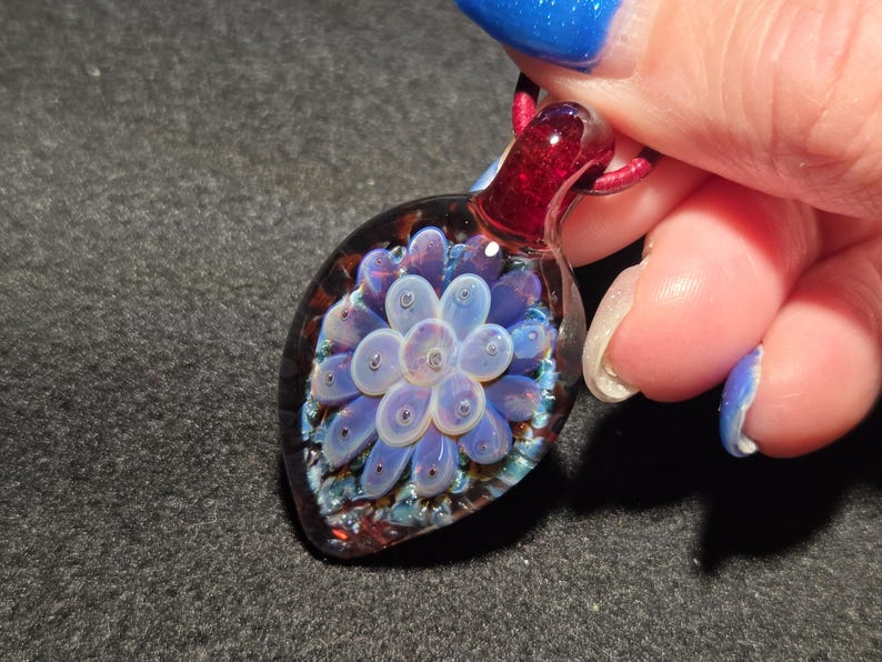 Hand-blown borosilicate glass implosion pendant featuring a pale lavender floral design encased in clear glass, set against a deep crimson red background with subtle blue sparkle accents, shown close-up against a dark surface.