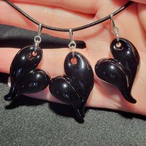 Three black glass heart shaped pendants hanging from a black leather cord. The pendants are all different shapes and sizes. The pendants are shiny and have a smooth surface.