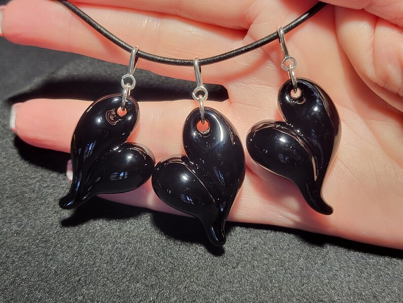 Three black glass heart shaped pendants hanging from a black leather cord. The pendants are all different shapes and sizes. The pendants are shiny and have a smooth surface.
