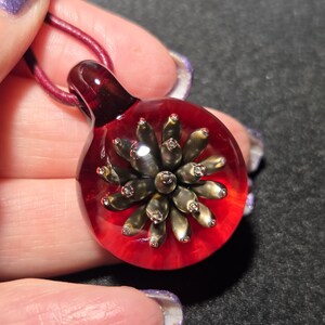 Close-up of a round glass pendant with a striking bronze-gold petaled implosion encased in clear glass and backed by a vivid transparent red, displayed in the palm of a hand.
