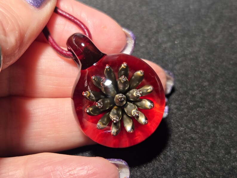 Close-up of a round glass pendant with a striking bronze-gold petaled implosion encased in clear glass and backed by a vivid transparent red, displayed in the palm of a hand.