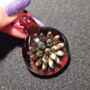 A borosilicate glass pendant with a metallic bronze and gold flower implosion encased in clear glass and set against a glowing red background, displayed on a black surface.