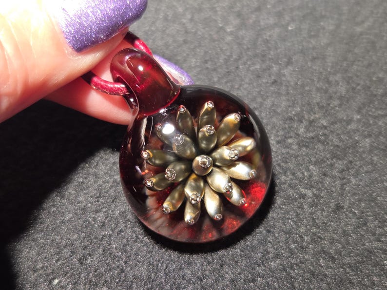 A borosilicate glass pendant with a metallic bronze and gold flower implosion encased in clear glass and set against a glowing red background, displayed on a black surface.