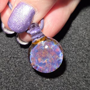 Sparkly handmade borosilicate pendant with a colorful frit implosion, purple glass bail and luminous blue, pink and gold details against black.