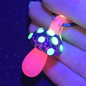 UV-reactive borosilicate glass mushroom pendant glowing fluorescent green and pink under blacklight, showing dramatic color transformation from its natural blue tones.